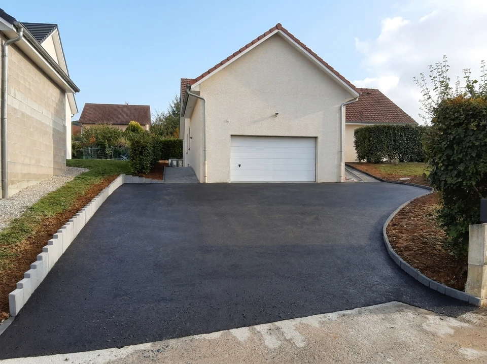 Allée de garage neuve en enrobé bitumineux noir, fraîchement posé, desservant un garage individuel avec porte sectionnelle blanche